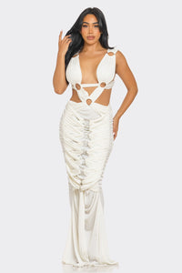 Sahara Mystique Pleated Gown - Model wearing a stunning beige cutout maxi dress featuring a plunging neckline, circular ring accents, and intricate ruched detailing throughout. The floor-length design with a figure-hugging silhouette exudes elegance and bold sophistication, perfect for an upscale evening event. - Rêves Dorés