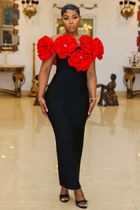 Fleur de Luxe
A confident woman stands in an elegant, vintage-inspired setting, wearing a refined black gown with eye-catching, oversized red floral embellishments along the neckline. The figure-hugging dress is paired with simple yet sophisticated black strappy heels. Her hair is sleek and stylishly arranged, and she accessorizes with a delicate bracelet and a gold watch. FEMME DE LUXE MUURSWAGG X ATHINA COLLECTION